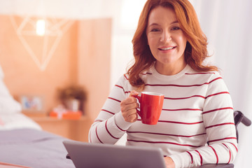Best beverage. Cheerful disabled woman using her laptop while drinking coffee