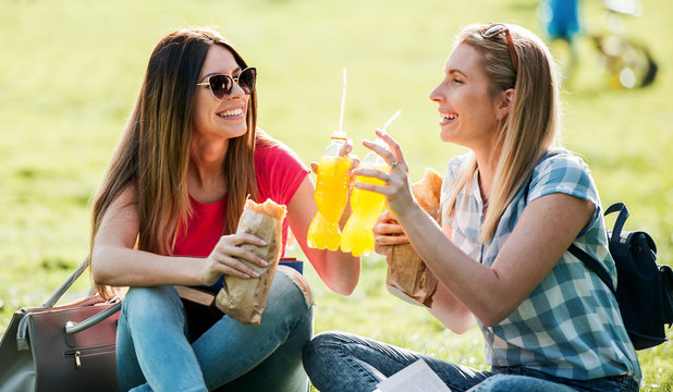 Campus life. Two young girls sitting in the park, eating sandwiches and drinking juice. Lifestyle concept