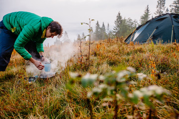 young man in green down jacket are heated in a fire and cooking hot drink out on a autumn camp in...