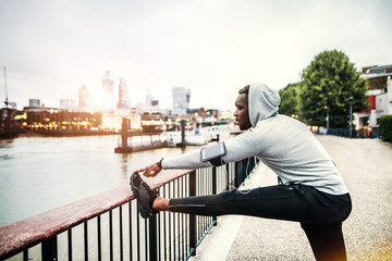 Black man runner with smartphone in an armband on the bridge in a city, stretching.