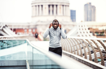 Black man runner with smartphone in an armband on the bridge in a city, resting.