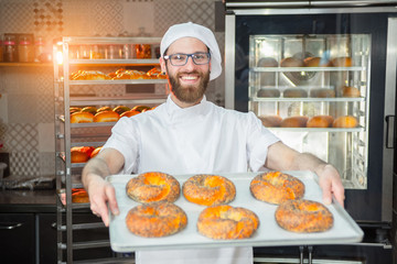 A young handsome baker holding fresh bagels with poppy seeds on a tray on the background of an oven and a rack with baked goods