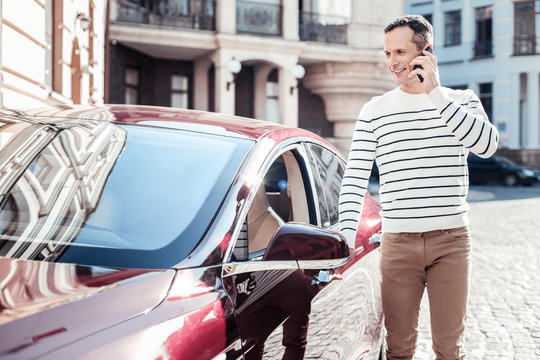 Favorite place. Smiling male person holding telephone near his ear and turning head while standing near his auto