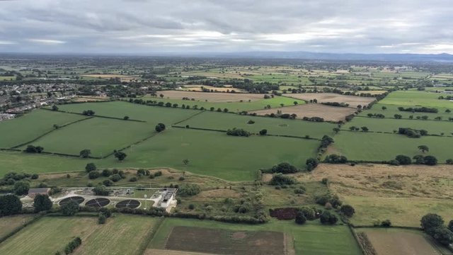 Aerial view, panning move. Sewage treatment plant, houses among fields on Cheshire countryside. Merseyside in background
