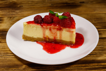 Piece of tasty New York cheesecake with raspberries and raspberry jam in a white plate on wooden table