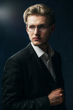Waist up studio portrait of young confident handsome man, businessman wearing round glasses, classic white shirt, black jacket, posing on dark background