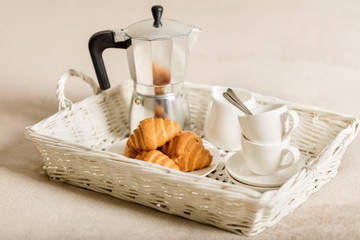 Breakfast in bed. On a white wicker tray there is a coffee maker, coffee white cups and croissants.