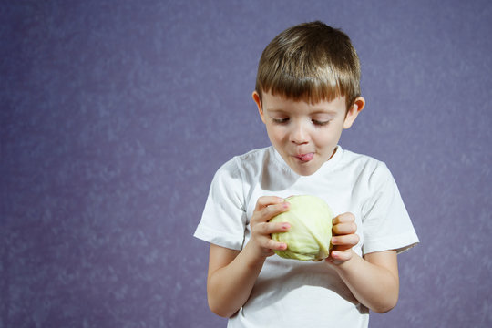 A small boy in a white T-shirt on a purple background holds a cabbage cabbage and lickens