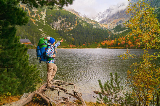 Beautiful young woman with big tourist backpack wearing blue outdoor jacket and knitted hat near chalet hotel. Lake Popradske Pleso in High Tatras Mountains. Start popular hiking ways.