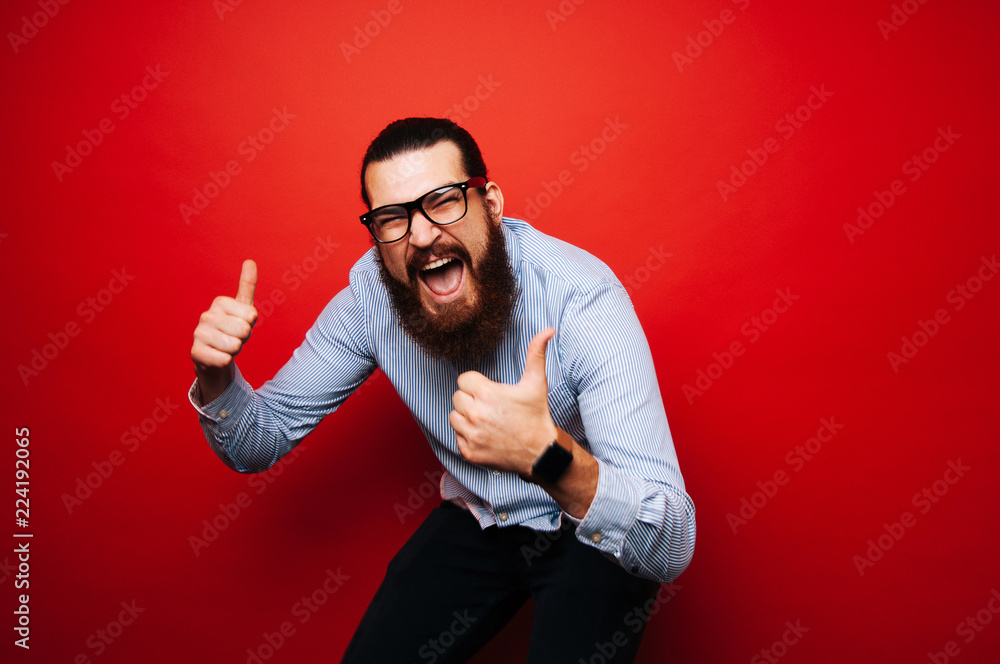 Wall mural cheerful bearded hipster man in casual with eyeglasses jump over red background and showing thumbs u