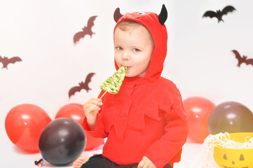 Fototapeta na wymiar Tricky cute boy in devil costume, sitting on a background of decorations for Halloween and eating green lollipop.