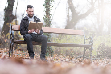 A handsome young hipster man sitting on a bench and reading a book in the park.