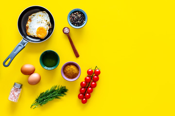 Recipe of fried eggs with vegetables. Ready eggs in a frying pan near cherry tomatoes, greenery, spices, raw eggs on yellow background top view copy space