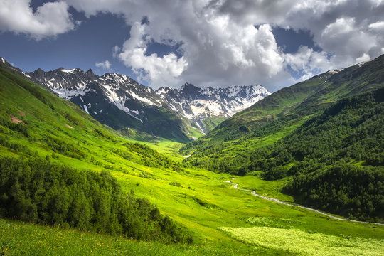 Alpine mountains landscape on bright sunny summer day. Grassy meadow on hillside with mountain river and rocky mountain covered by snow. Blue sky with clouds over mountain range. vibrant highlands