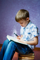 Fototapeta premium Little boy in a light shirt sits on a pile of books with a green book in his hands and looks through a magnifying glass