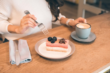 cropped image of young woman with coffee cup eating cheesecake at table in cafe