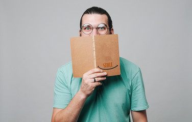 Young hipster wearing glasses looking at the camera an hiding part of face behind book. Smile word.