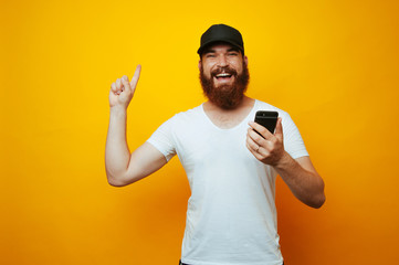 Happy bearded man holding phone and pointing up on yellow background.