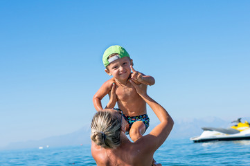 Young mother in black sunglasses and smiling baby boy son in the green baseball cap playing in the sea in the day time. Positive human emotions, feelings, joy.