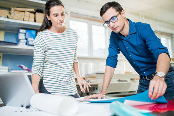 Young casual man and woman in printing office working in team and choosing papers and colors at table