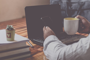 A Man are using the laptop computer to working and Holding a mug of coffee. Have a pen and paper and on a wooden table. Image is Mood tone.