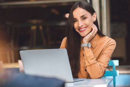 smiling young female freelancer looking at camera while working on laptop at table in cafe at urban street