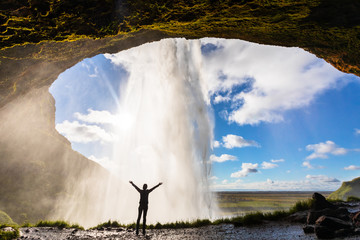 Happy female traveler feeling the power of Seljalandsfoss waterfall in the South of Iceland, person standing behind the stream, famous Icelandic landmark