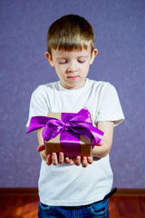 A little boy in a white T-shirt on a purple background holds a gift with a big bow and is sad