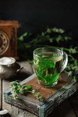 A cup of hot herbal tea made from lemon balm on a wooden background. Melissa branches, sugar bowl,...
