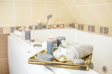 Fototapeta na wymiar Candles burning and spa essentials on wooden tray in bathroom, essential oil, bath salt on wooden spoon, pumice stone, towel, sea stone, white orchids. Spa products set conception. 