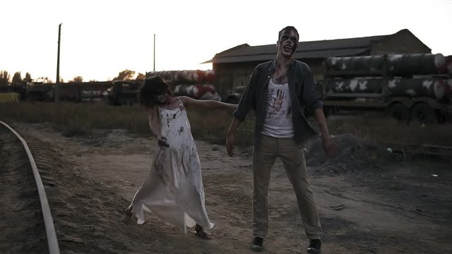 Creepy two zombies in bloody clothes walking through the ruined city during the zombie apocalypse. Abandoned place with trucks with missiles on the background. Front view