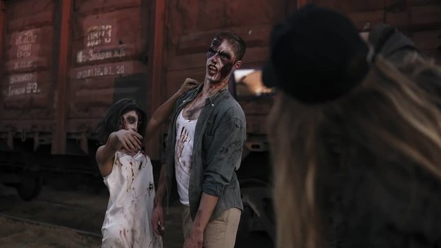 The girl from the backstage making a photo with her mobile of two zombies standing in front the wagon outdoors. Horror, halloween, staging concept