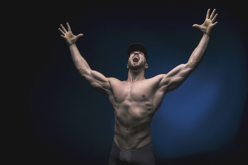 Fototapeta na wymiar Young strong man bodybuilder in cap on wall background. Dark dramatic colors.