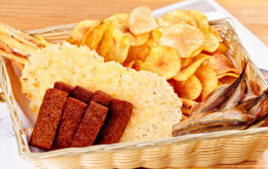 Assorted snacks for beer: sun dried fish, potato chips, salted crackers, rye bread croutons in basket on wooden background