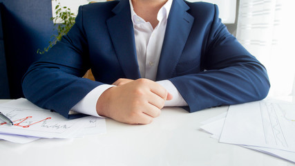 Closeup image of businessman in blue suit sitting behind desk covered with graphs and charts