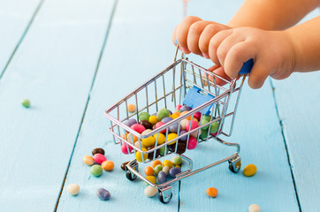 Hands of a child holding a shopping cart with colored sweets on a blue background