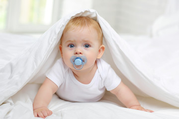 Charming blue-eyed baby 7 month old lies in bed in a white bodysuit, covered with a blanket and sucks a pacifier