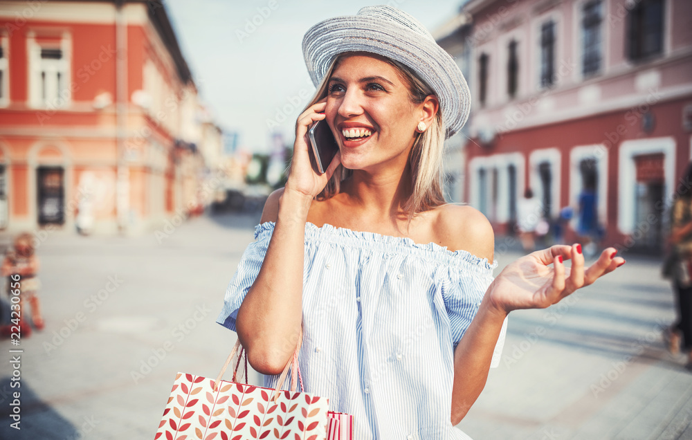 Wall mural Shopping time. Young woman talking on a mobile phone while walking down the street. Consumerism, shopping, lifestyle concept