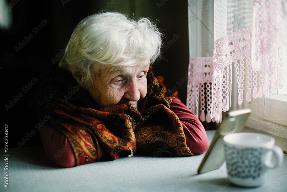 Wall mural Elderly woman sitting at the table looking at the screen of the smartphone.