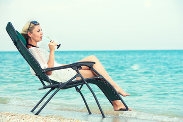 Young woman sits on the beach on a chaise longue and drinking wine.	