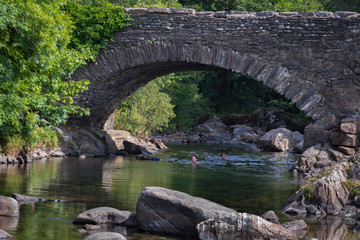 Fototapeta na wymiar Father and daughter swimming in the River Duddon by old stone bridge in Ulpha in the Lake District National Park, UK. Scenic view of English countryside on a sunny summer day.