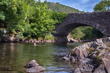 Teenage boy is swimming in the River Duddon by old stone bridge in Ulpha in the Lake District National Park, UK. Scenic view of English countryside on a sunny summer day.