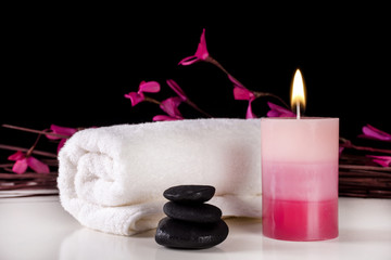 Aromatic candle burning on table and white towel and black stones, black background with purple flowers on branches. Spa treatment concept
