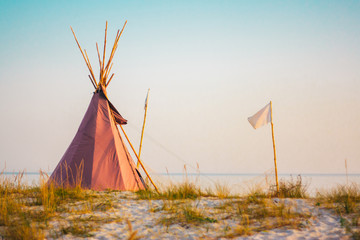 Red wigwam and white flag on the sandy seashore. Indian native national building outdoors summer....