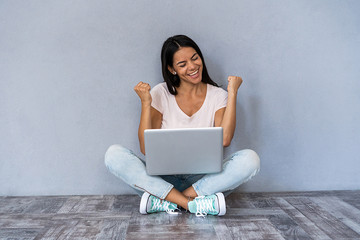 Portrait of excited young casual girl celebrating success while sitting with laptop computer...