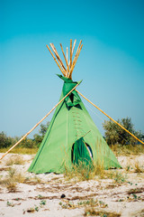 Green wigwam on the sandy seashore. Indian native national building outdoors summer. Teepee on coast on blue  sky background © baklykovadaria