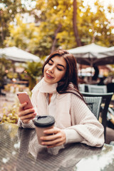 Beautiful young woman having coffee in outdoor cafe while using smartphone. Portrait of stylish girl typing message