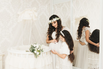 Attractive pregnant mom with her daughter are standing near the crib for . Photos of pregnancy in...