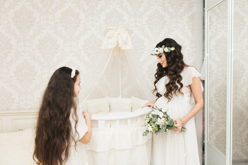 Attractive pregnant mom with her daughter are standing near the crib for . Photos of pregnancy in...