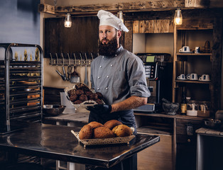 Handsome bearded chef in uniform showing tray of fresh bread in the kitchen of bakery.